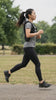 A woman running side-on through a park wearing a black HEFT weighted vest over a grey tee